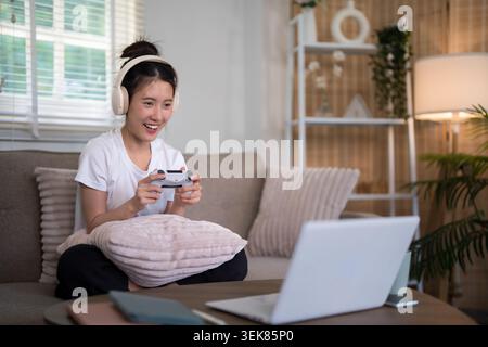 happy young Asian woman wearing headphones playing video games using game controller at home on couch indoor living Stock Photo
