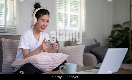 Asian woman play video game with headphones and controller relax on sofa at home casual leisure entertainment lifestyle Stock Photo
