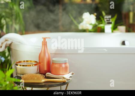 Bathroom scene with white bathtub, coral bath products, candle, & wooden hairbrush on side table. Blurred green plants & dark tiles enhance tranquil, Stock Photo