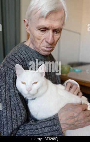 Senior man holding white cat in living room at home. Elderly pet owner supporting feline with warmth and gentle affection. Grandfather cradling beloved animal showing mature bonding and closeness Stock Photo