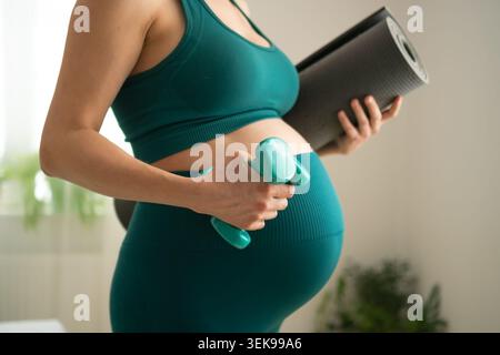 Expecting mother standing with dumbbell for active strength training in pregnancy. Pregnant woman showing determination to stay fit. Mom-to-be combining light weights for prenatal fitness routine Stock Photo