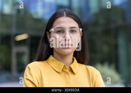 Young business woman wearing a yellow shirt and clear eyeglasses, standing outdoors in an urban setting, representing professionalism, intelligence, and a serious determined attitude for her career Stock Photo