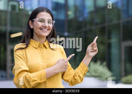Smiling businesswoman in yellow shirt and clear glasses pointing both hands to the side, presenting or showing important information or directions outside a modern office building Stock Photo