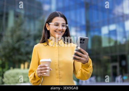 Young indian businesswoman smiling while looking at her smartphone and holding a takeaway coffee cup outdoors in an urban environment with modern buildings Stock Photo