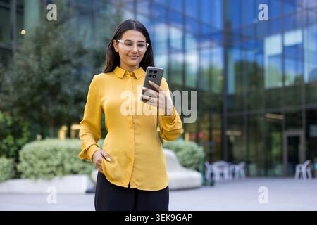 Professional woman in yellow blouse and clear glasses checking her smartphone, standing in front of a modern glass building, utilizing mobile technology for communication Stock Photo