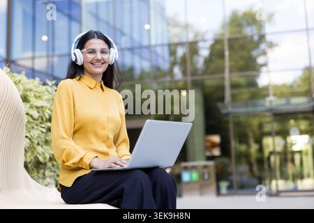 Young woman smiling, wearing headphones and clear glasses, working or studying on a laptop while sitting on a modern bench outdoors in front of a glass building Stock Photo