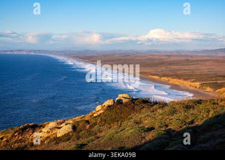 Rolling waves crashing along a vast sandy beach and rugged coastline at sunset Stock Photo