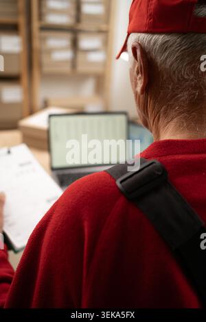 Senior elderly postal worker using a digital tablet to check parcel boxes in a post office, wearing a overalls and cap Stock Photo