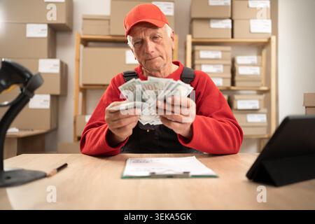 Elderly postal worker in uniform holding cash money near parcels at post office. Concept of payment, profit, salary, financial success, lottery win Stock Photo
