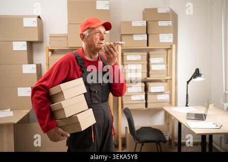 Elderly postal worker holding parcels in warehouse using AI voice assistant Stock Photo