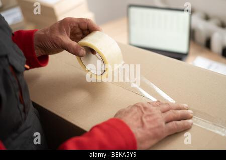 Close up of elderly postal worker hands packing cardboard parcel box for shipment at post office. Senior employee preparing package for delivery Stock Photo