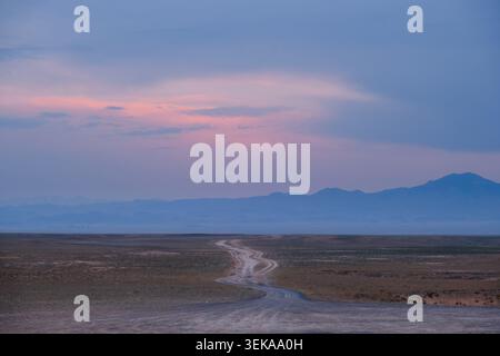 Winding dirt road running through a vast desert landscape toward distant mountains under a twilight sky with pink clouds. Ideal commercial background Stock Photo