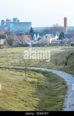 A quiet rural village scene featuring a curved gravel path through a grassy meadow, residential houses, a brick tower and distant industrial silos und Stock Photo