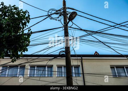 A utility pole in Tirana, Albania, tangled with a dense, chaotic mess of overhead power lines and cables against a building and blue sky. Stock Photo