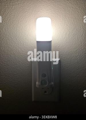 LED Night Light Plugged Into Electrical Wall Outlet Illuminating Dark Interior Wall Stock Photo