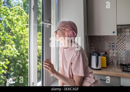 Middle aged woman standing by open window at home, breathing fresh air, experiencing relief after stuffy indoor air. Stress relief, indoor air quality Stock Photo
