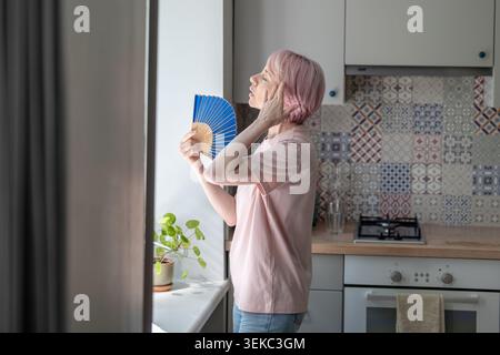 Middle aged woman standing by window holding paper hand fan, feeling tired from hot weather and stuffy indoor air. Concept of summer heat, heat stress Stock Photo