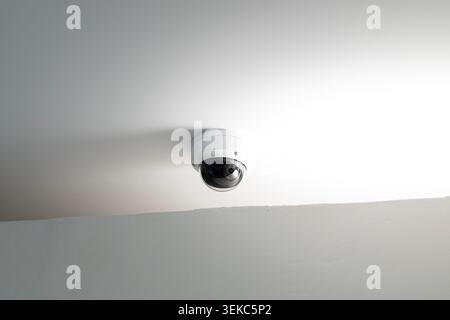 Sharp and detailed view of a white dome security camera mounted on a plain white ceiling. Strong, bright light from the side creates a high contrast r Stock Photo