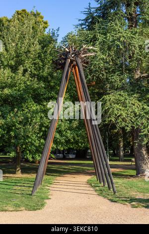 Tall wooden sculpture stands beside a park path among mature trees and grass in Annecy, France, during a sunny summer afternoon. Stock Photo