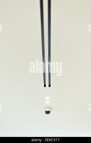 Sharp view looking up at a white minimalist ceiling featuring a dome security camera, two small recessed spotlights, and parallel black track lighting Stock Photo