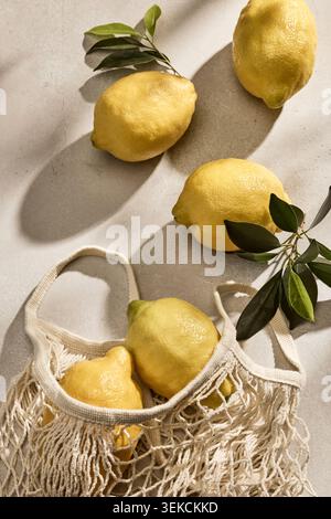 Overhead flat lay shot of fresh, vibrant yellow Italian lemons inside a reusable eco-friendly woven mesh string bag. The bag rests on a kitchen table Stock Photo