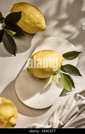 Overhead flat lay shot of three vibrant fresh Italian lemons with natural green leaves attached, resting on a rustic kitchen tabletop surface. Capture Stock Photo
