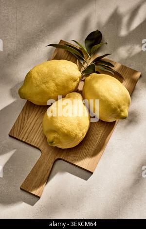 Overhead flat lay shot of three vibrant fresh Italian lemons with natural green leaves attached, resting on a rustic kitchen tabletop surface on woode Stock Photo