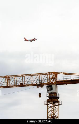 Passenger airplane passes above a yellow tower crane at an active urban construction site under cloudy sky. Stock Photo