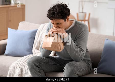 Young man with paper bag having panic attack on sofa at home Stock Photo