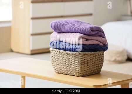 Wicker basket with stack of knitted sweaters on bedside bench in bedroom, closeup Stock Photo