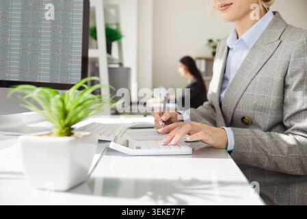 Businesswoman using calculator and taking notes while analyzing financial data on computer screen. Stock Photo