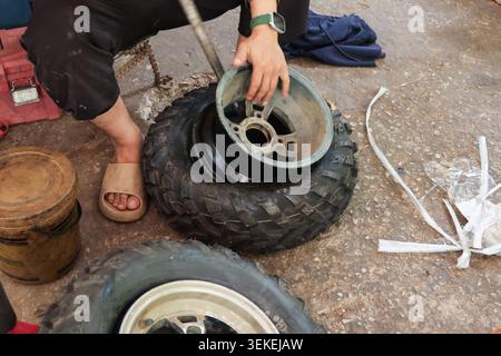 Mechanic repairing flat tire on rim with metal tool sitting on ground near bucket concentrating on manual labor outdoors in casual clothing and sandal Stock Photo
