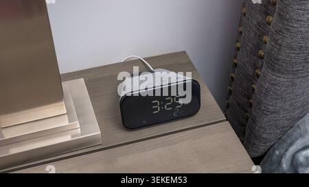 Smart clock with digital display showing time on a wooden bedside table next to a lamp and textured fabric headboard in a modern bedroom setting Stock Photo