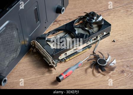 Disassembled graphics card, fan blade, screwdriver, and screw lie on wooden table near open PC case. Concept of computer repair, cleaning, and hardware maintenance. Stock Photo