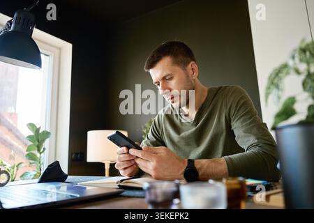 Focused man using smartphone at desk beside laptop and open notebook. Concept of planning, organization and productive work at home. Stock Photo