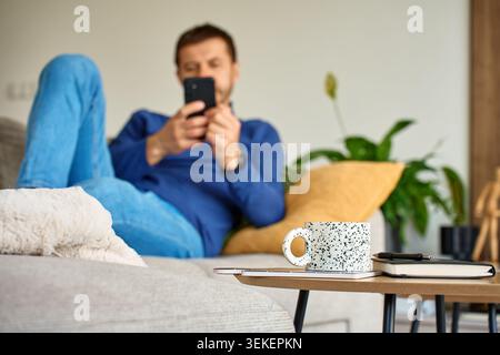 Adult man lying on sofa and uses smartphone in living room with mug and notebook on table. Concept of home lifestyle, mobile technology and everyday leisure Stock Photo