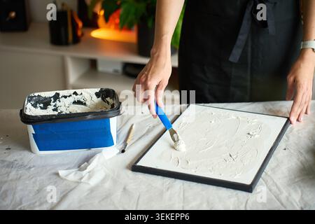 Artist making textured canvas art. Woman applying white plaster paste with a blue palette knife at table covered with protective sheet at home. Concept of handmade craft and creative hobby Stock Photo
