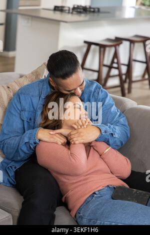 Mid-adult couple sitting on light-grey sofa in living room, embracing with smartphone on lap Stock Photo