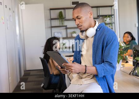 Diverse coworkers working in office, man sitting on desk holding tablet wearing headphones Stock Photo