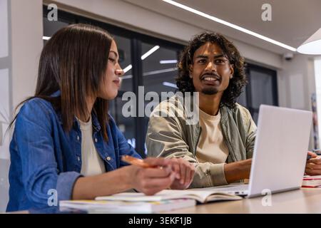 Asian woman and Indian man writing with pencil by notebooks and laptop at modern study table Stock Photo