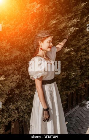 Woman fashion sunset smiling elegant lady in white puff sleeve dress touching green foliage in garden during golden hour light Stock Photo