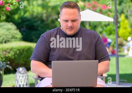 Young man concentrating while working on a laptop outdoors in a sunny green garden. Authentic lifestyle and business image representing remote work, d Stock Photo