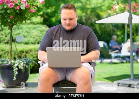 Young man working on a laptop outdoors in a sunny green garden. Authentic lifestyle and business image representing remote work, digital nomad lifesty Stock Photo