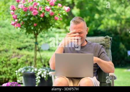 Exhausted mature man sitting outdoors with a laptop and reacting with frustration and stress. Concept for technology overload, online problems, remote Stock Photo