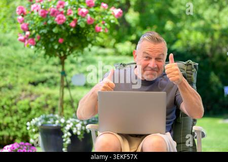 Mature man sitting outdoors with a laptop and giving thumbs up gesture in a sunny garden. Concept for customer satisfaction, positive online experienc Stock Photo