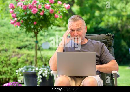 Mature man reacting with shock and concern while using a laptop outdoors in a sunny garden. Authentic lifestyle image illustrating unexpected online p Stock Photo