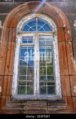 Old arched window with peeling white paint set in red brick wall. Glass panes show reflections of greenery and sky. Stone frame weathered from time an Stock Photo