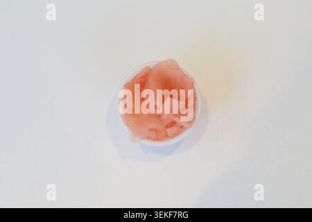 Pickled ginger slices in a small plastic pot on a white background for sushi, poke bowls and Asian meals. Stock Photo