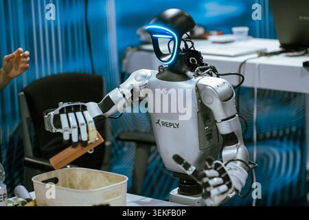 A humanoid robot with a glowing blue visor interfaces with an object over a container during a live automation and programming demonstration at a tech Stock Photo