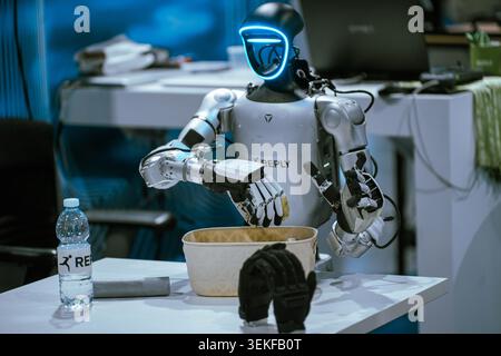 A humanoid robot with a glowing blue visor interfaces with an object over a container during a live automation and programming demonstration at a tech Stock Photo